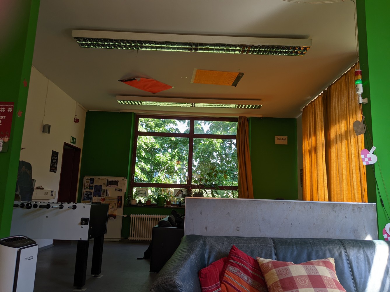 Blick in die Lounge: links Tuschkicker,im Vordergrund ein Sofa mit roten Kissen, grüne Wände und im Sonnenlich orange Vorhänge. Im hintergrund Fensterfront mit Blick ins Grüne.