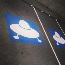 Foto eines Parkplatzes. Auf dem Boden eine blaue Markierung mit den weißen Umrissen eines Ufos.