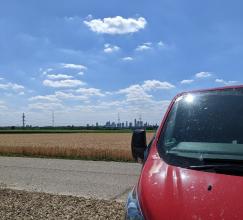 Symbolbild Ausflug: rotes  Auto im Vordergrund vor Kornfeld, blauem Himmel und weißen Wolken. 